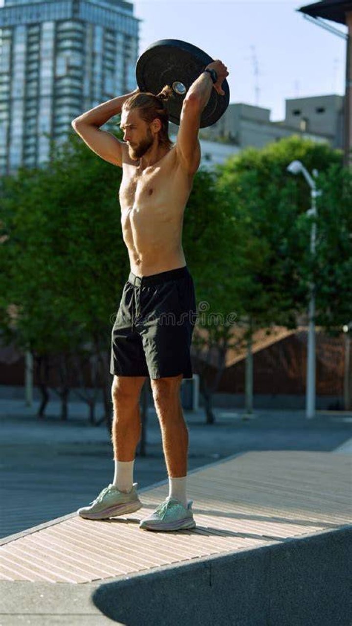 young shirtless muscular man training with weights outdoors at the sunset